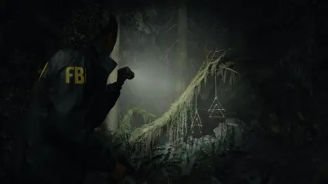 Alan Wake 2 – Screenshot von Saga Anderson, die mit ihrer Taschenlampe einige okkult aussehende dreieckige Symbole anleuchtet, die im Wald von einem Ast herunterhängen