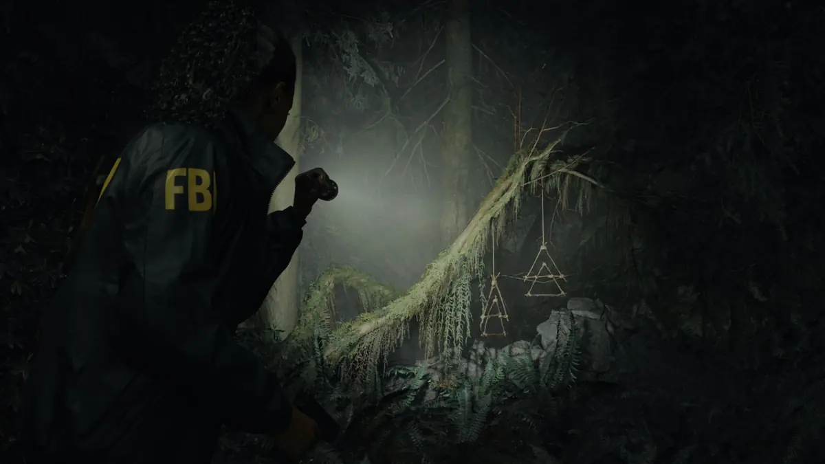 Alan Wake 2 – Screenshot von Saga Anderson, die mit ihrer Taschenlampe einige okkult aussehende dreieckige Symbole anleuchtet, die im Wald von einem Ast herunterhängen